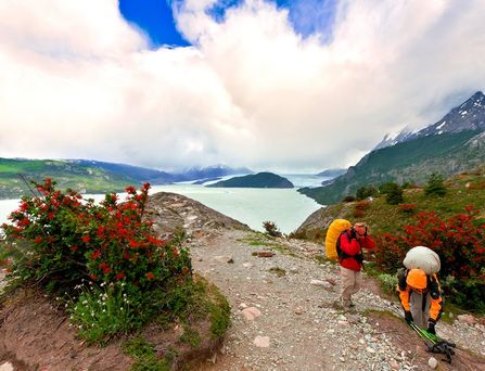 Zwei Wanderer mit großen Rucksäcken auf einem felsigen Pfad, umgeben von roten Beerensträuchern, mit Bergsee und schneebedeckten Bergen im Hintergrund.