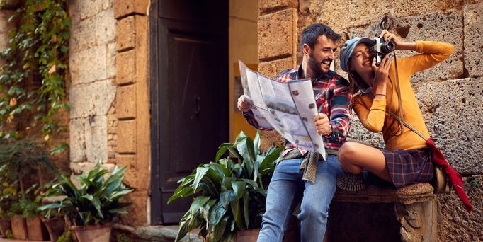 Ein junges Paar sitzt in einer mediterranen Gasse auf einer Steinbank. Er hält einen Stadtplan in der Hand und blickt lachend zu seiner Freundin. Sie lächelt ebenfalls und hält die Kamera vor ihr Gesicht um ein Foto zu schießen.