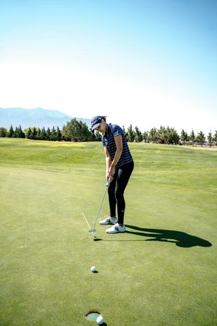 Eine Person in Golfkleidung und Cap steht auf einem grünen Golfplatz und hat gerade gegen den Golfball geschlagen, welcher kurz vor dem Loch liegt.