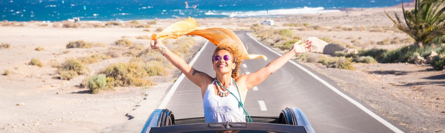 Eine sommerlich, hippimäßig gekleidete Frau sitzt in einem Cabrio mit offenem Dach. Sie steht im Auto und reist die Arme lachend nach oben und ein orangenes Tuch weht im Wind. Im Hintergrund das wilde Meer.