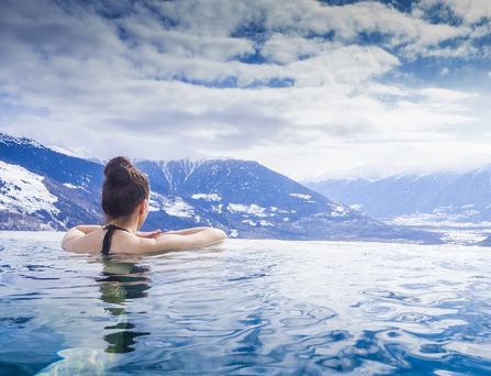 Eine Frau mit braunem Dutt lehnt am Rand eines Infinitypools mit Blick auf die verschneiten Bergspitzen.