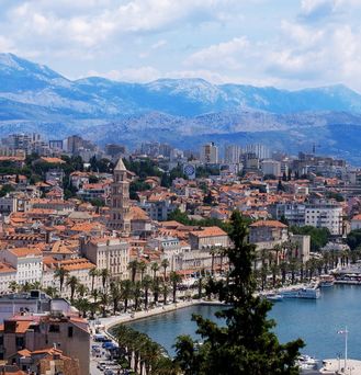 Die Hafenpromenade von Split mit Palmen am Ufer, mediterranen Häusern mit orangenen Dächern und Bergen im Hintergrund.