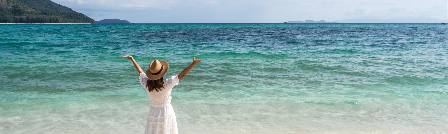 Eine Frau mit weißem Sommerkleid und Sonnenhut steht vor dem Meer und reißt die Arme freudig in die Luft.
