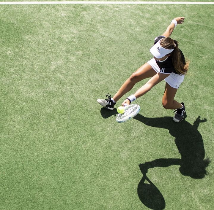 Eine braungebrannte junge Frau in Tenniskleidung befindet sich auf einem Tennisplatz und spielt gerade den Ball zurück, während die Sonne einen schwarzen Schatten auf den grünen Platz wirft.