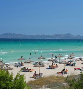Ein von Sonnenliegen mit Sonnenschirmen geprägter Strand. Im Hintergrund sieht man Berge