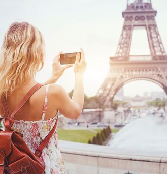 Eine junge, sommerliche gekleidete Frau steht vor dem Pariser Eifelturm und schießt mit ihrem Handy ein Foto im Querformat.