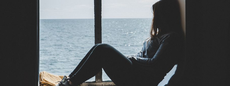 Eine Frau hockt auf der Fensterbank eines großen Schiffsfensters und schaut raus auf das offene Meer.