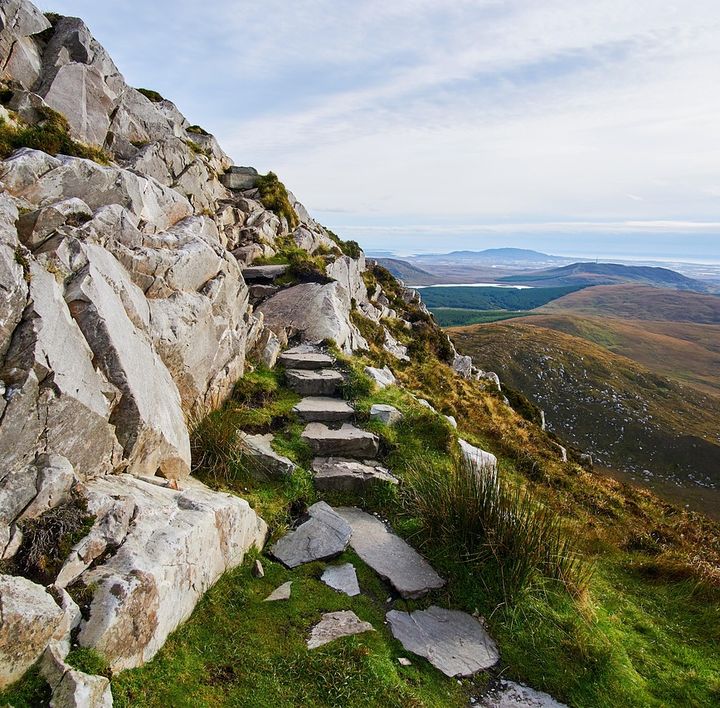 Eine Treppe aus Steinen führt an einer felsigen Mauer hoch. Rechts am Abgrund das Landschaft der Highlands.