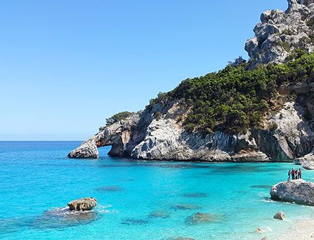 Kristallklares, türkises Wasser an einer steinigen, steilen Bucht. Ein paar Menschen stehen auf einem Felsen.