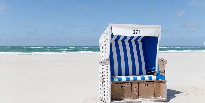 Ein blau-weiß gestreifter Strandkorb steht am weißen Sandstrand.