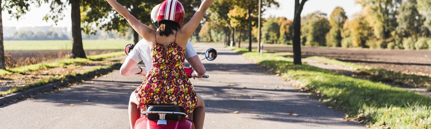 Ein Mann und eine Frau sitzen in sommerlicher Kleidung auf einem roten Roller. Die Frau mit rotem Helm sitzt hinten und reißt beide Arme in die Luft. Die beiden fahren auf einer ruhigen Landstraße zwischen einer grünen Allee.