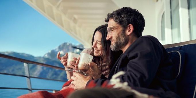 Ein Paar sitzt mit 2 Kaffeegetränken auf einem Schiffsbalkon und schaut lächelnd aufs Meer
