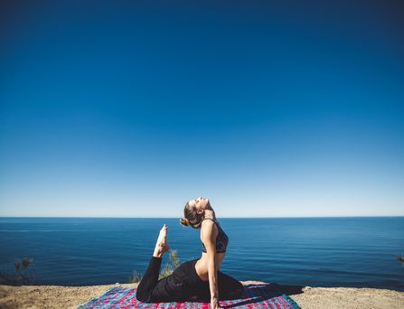 Eine Frau sitzt in einer Yoga-Stellung auf einer Decke an einem Abhang. Im Hintergrund das tiefblaue Meer oder ein See.