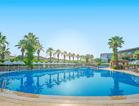 Um einen großen türkisen Pool stehen weiße mehrstöckige Häuser mit Palmen und Sonnenliegen davor.