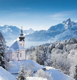 Eine kleine, weiß-rosa-farbene Kapelle ragt aus einer winterlichen Landschaft mit schneebedeckten Tannen und Berggipfeln hervor.