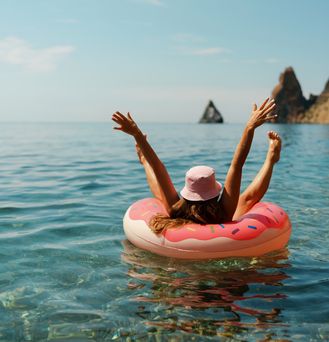 Ein Mädchen mit Sommerhut liegt in einem pinken Gummidonut auf dem Meer. Sie reißt Beine und Arme in die Höhe.