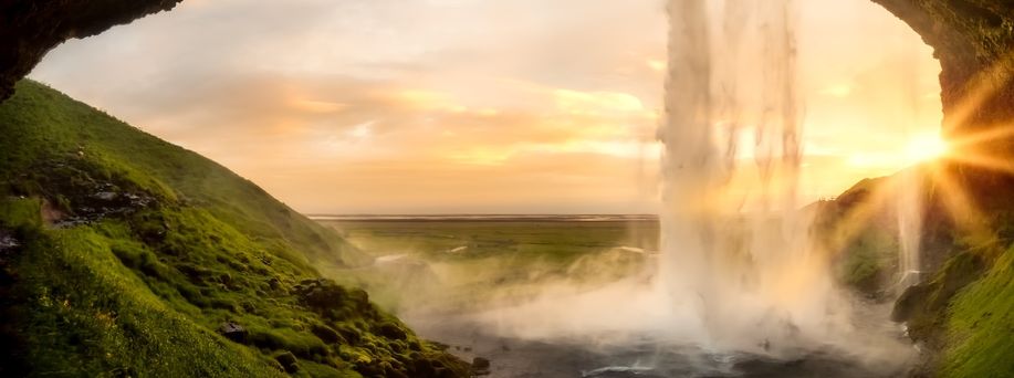 Aus einer Felshöhle heraus führt der Blick in eine weite grüne Landschaft mit untergehender Sonne. Von oben führt ein kräftiger Wasserfall auf die schwarze Erde.