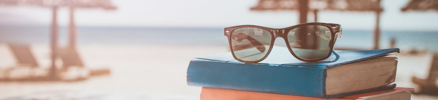 Auf 2 Büchern liegt eine Sonnenbrille und im Hintergrund ist leicht verschwommen ein Strand mit Liegestühlen und Sonnenschirmen.