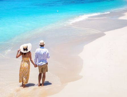 Reiseziele weltweit: Ein Paar mit Sonnenhüten läuft Hand-in-Hand an einem weißen Sandstrand mit türkisem Wasser entlang