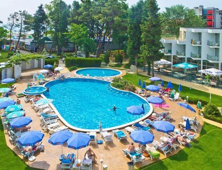 An einer verspiegelten Glaswand tropft ein künstlicher Wasserfall in einen türkisen Wassergraben der am Gebäuderand entlang führt. Daneben eine Wiese mit Blumen und Palmen.