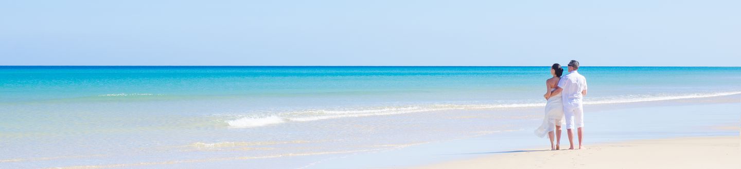 Ein Paar steht in weißer Sommerkleidung an einem weißen Sandstrand und blickt auf das kristallklare türkise Meer.
