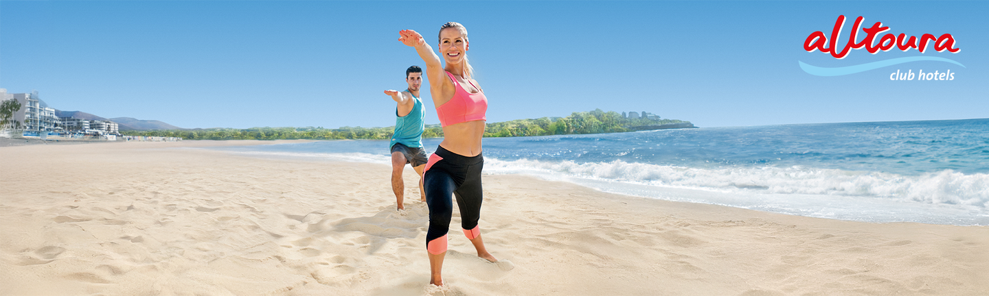 An einem hellen Sandstrand stehen eine Frau und ein Mann zwischen 25 und 35 in einer Yogapose und Sportklamotten und lächeln.