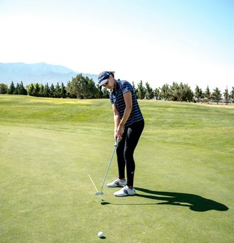 Eine Person in Golfkleidung und Cap steht auf einem grünen Golfplatz und hat gerade gegen den Golfball geschlagen, welcher kurz vor dem Loch liegt.