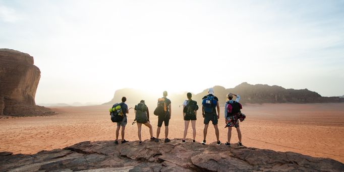 6 junge Menschen stehen mit Reiserucksäcken mit dem Rücken zugewandt auf einem Felsabsprung und blicken in die weite Landschaft. Im Hintergrund sind weitere braune Felsen zu sehen.