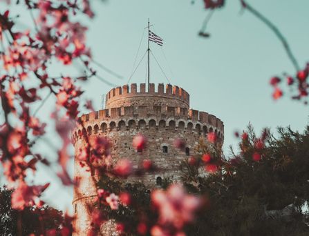 Eine mittelalterlicher Burgturm mit Griechenlandflagge ragt in den hellblauen Himmel. Im Vordergrund pinke Blumen.