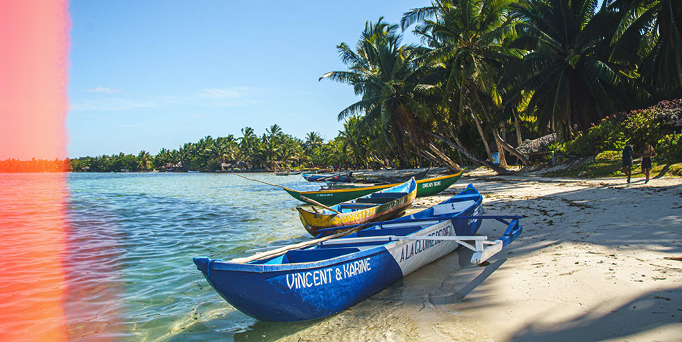 Mehrere bunte Fischerboote am sandigen Strand unter Palmen, rechts oben das Logo von Hapag-Lloyd Cruises.