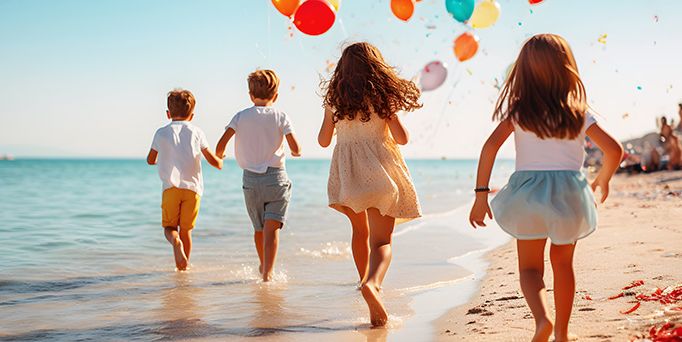 Vier Kinder in sommerlicher Kleidung und bunten, schwebenden Luftballons laufen am Strand entlang