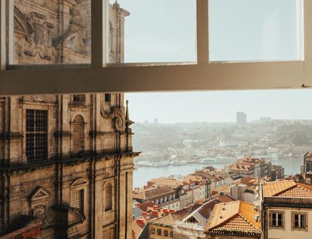 Blick aus einem Fenster auf ein imposantes, historisches Gebäude und eine Stadt mit Hafen im orangenem Licht.