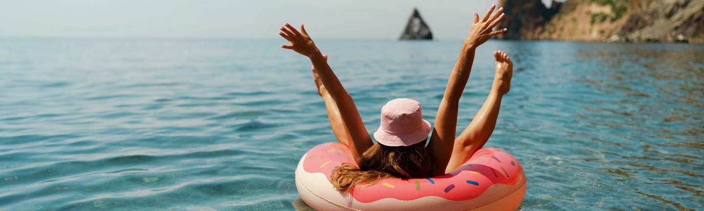 Eine Frau mit Sonnenhut liegt auf einer Luftmatratze, welche aussieht wie ein pinker Donut, auf dem Wasser. Sie streckt Beine und Arme in die Luft.