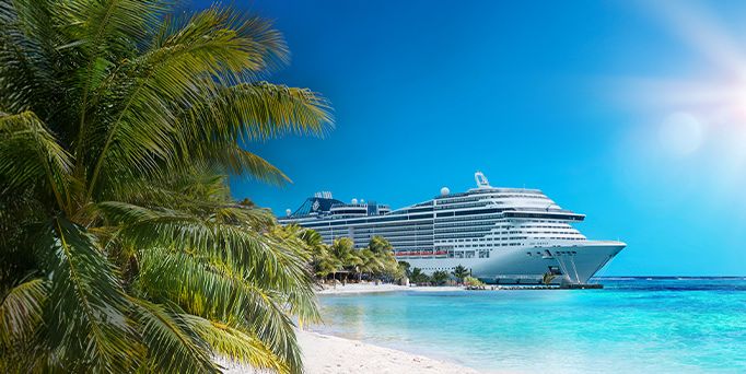 An einem karibischen Strand mit weißen Sand und türkisem Wasser stehen Palmen und im Hintergrund liegt ein riesiges weißes Kreuzfahrtschiff an. Das Logo der Reederei MSC befindet sich unten links im Bild. 