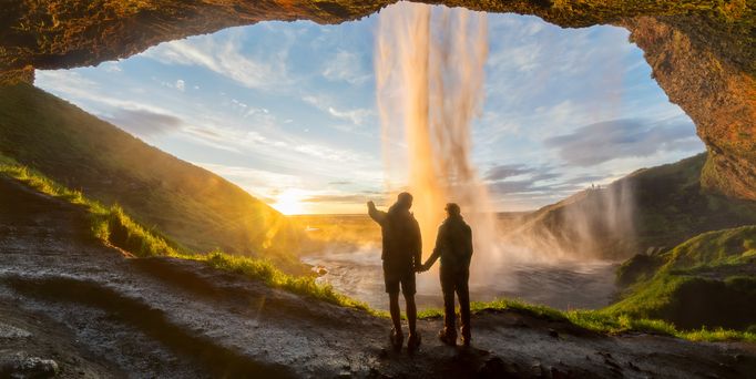 Aus einer Felshöhle heraus führt der Blick in eine weite grüne Landschaft mit untergehender Sonne. Von oben führt ein kräftiger Wasserfall auf die schwarze Erde.