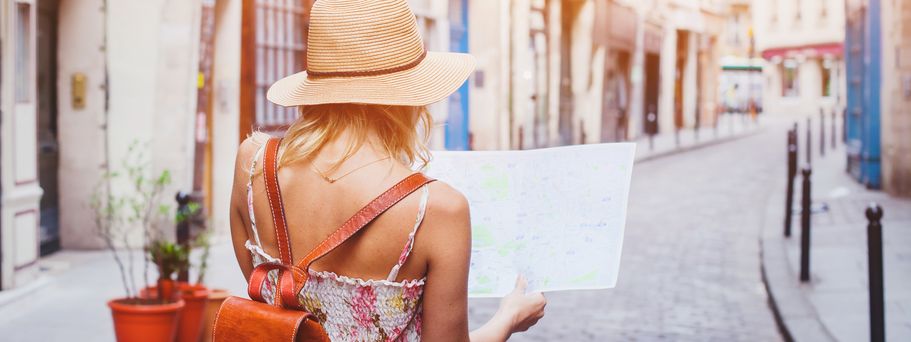 Eine junge Frau in Sommerkleidung, Sommerhut und mit Rucksack läuft mit einer Karte in den Händen durch eine mediterrane Gasse.