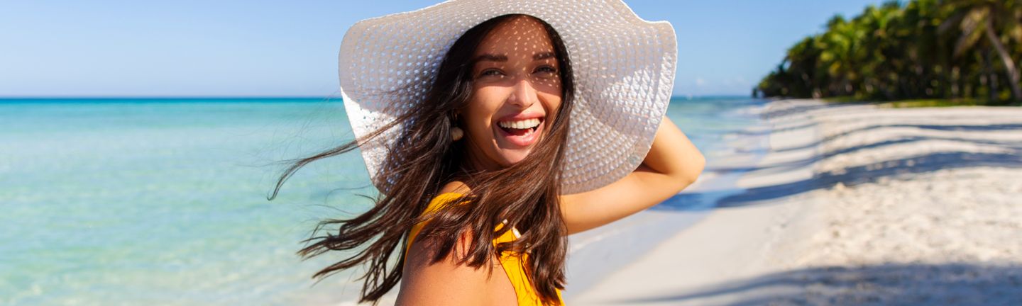 Reisetrends 2026: Eine Frau mit großem, weißen Sonnenhut läuft lachend an einem paradiesischen Strand entlang.