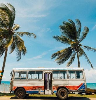 An einer Strandpromenade steht ein alter, rustikaler bunter Bus. Daneben und dahinter Kokospalmen.