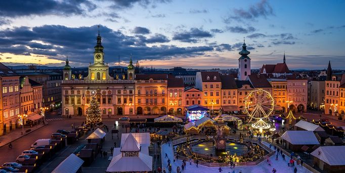 In der Abenddämmerung befindet sich ein Marktplatz umgeben von hübschen, Altbauten. Auf dem Platz ist ein beleuchtetes Riesenrad, Karussel und weihnachtliche Hütten.