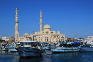 An einem Hafen legen mehrere rustikale, bunte Boote an. Im Hintergrund eine beige Moschee.