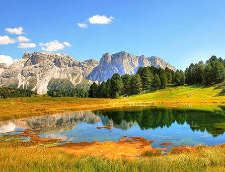 Eine imposante, von Schnee bedeckte Bergkette, spiegelt sich in einer riesigen Pfütze wieder. Diese liegt in einer gold-gelben Graslandschaft.