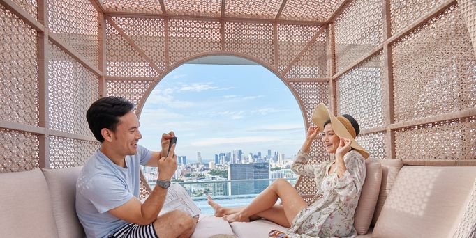 Ein asiatisches Pärchen sitzt in einer modernen Cabana und er schießt ein Foto von ihr. Durch ein rundes Loch ist die Skyline von Singapur zu sehen.