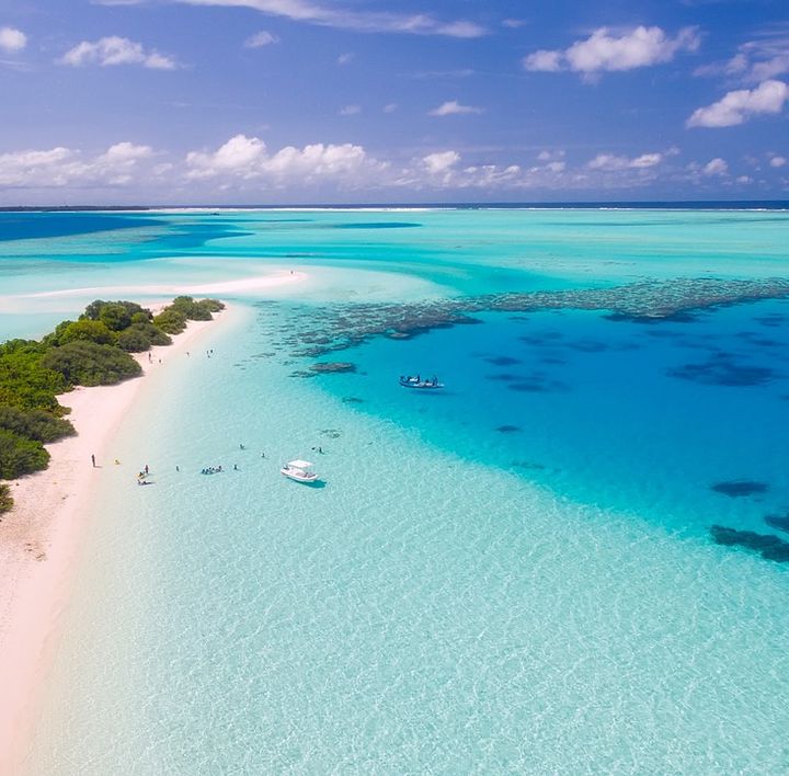 Ein schmaler weißer Sandstrand mit tropischr Vegetation, direkt am türkis, blauen Wasser mit zwei Booten am Ufer und einigen Menschen.