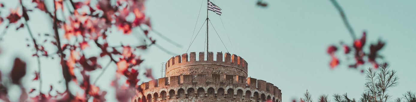 Eine mittelalterlicher Burgturm mit Griechenlandflagge ragt in den hellblauen Himmel. Im Vordergrund pinke Blumen.