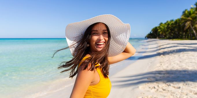 Reisetrends 2026: Eine Frau mit großem, weißen Sonnenhut läuft lachend an einem paradiesischen Strand entlang.