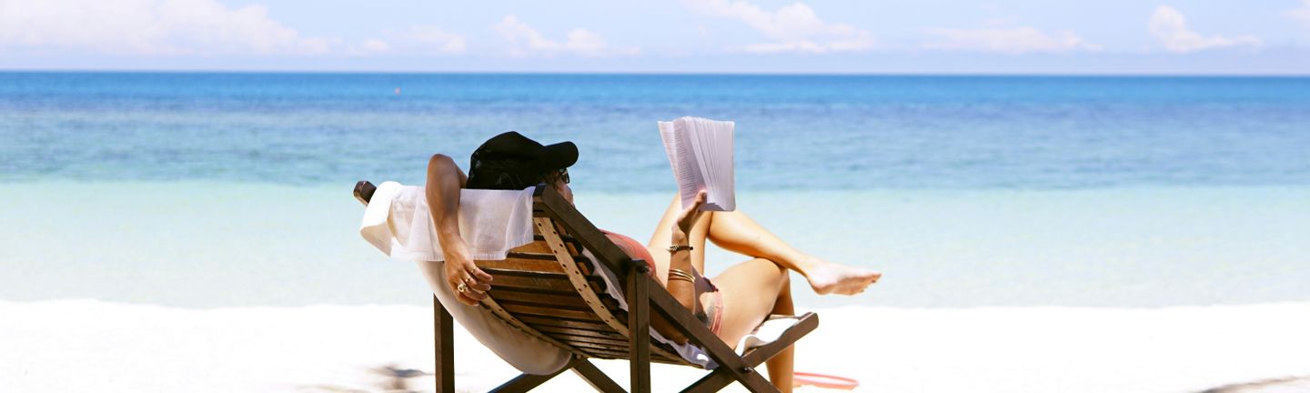 Eine junge Frau liegt entspannt mit einem Buch am Strand im Liegestuhl.