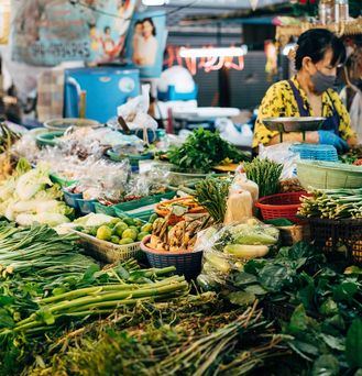 Eine grün-bunte Vielfalt an asiatischen Lebensmitteln, vor allem Gemüse liegt nebeneinander und teilweise übereinander auf einem Tisch auf einem Marktplatz. Hinter der Theke steht eine asiatische Frau mit Atemmaske.