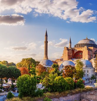Sicht auf eine imposante, schöne Moschee, mit mehreren Kuppeln, welche aus einem bunt gefärbten Blätterdach emporragt.