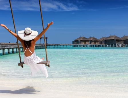 Malediven Urlaub: Eine Frau mit weiße Sommerkleid und Hut sitzt auf einer Schaukel am Strand.
