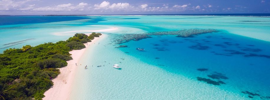 Ein schmaler weißer Sandstrand mit tropischr Vegetation, direkt am türkis, blauen Wasser mit zwei Booten am Ufer und einigen Menschen.